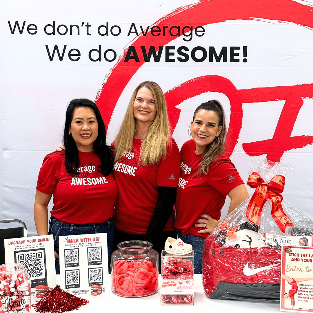 Three women in red shirts promoting DiCiccio Freeman Orthodontics at a community event, with a display of promotional materials, including QR codes and giveaways, emphasizing community engagement and orthodontic services in Fresno.
