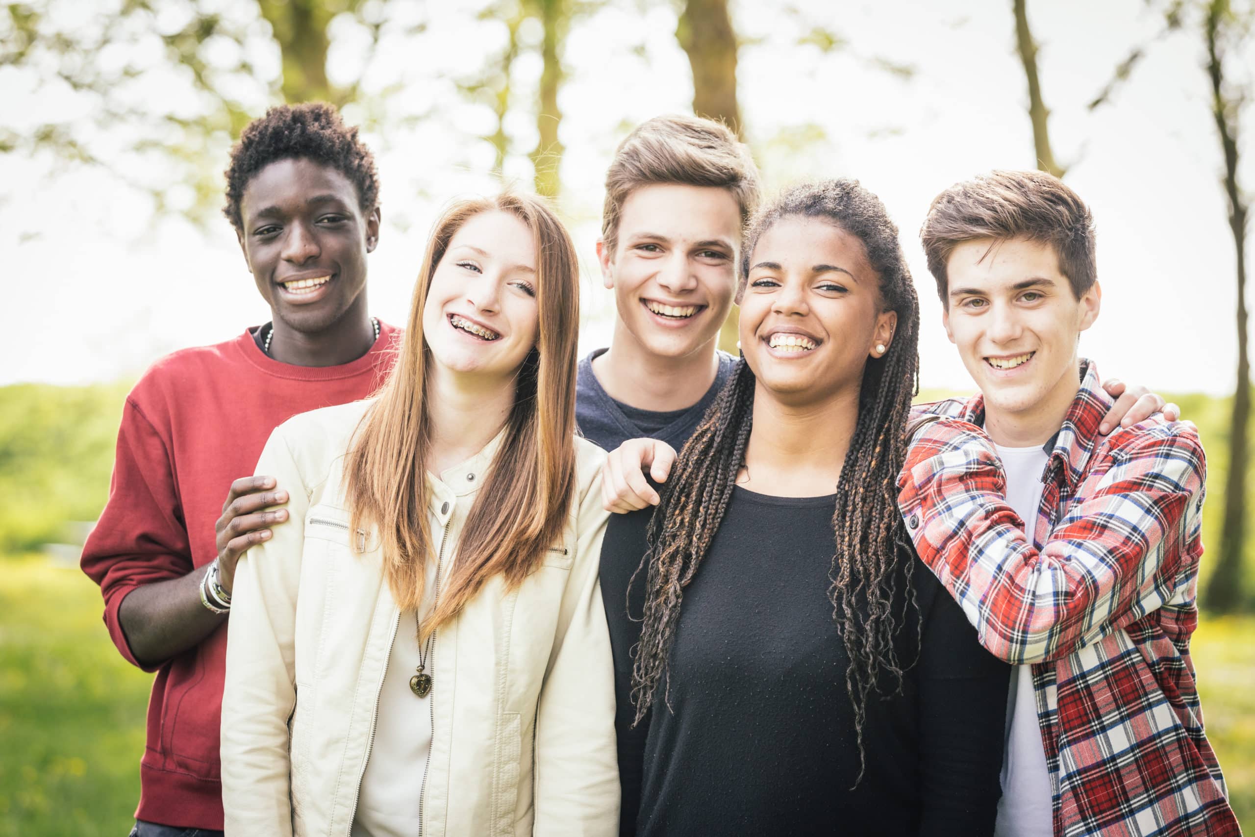 Multiethnic group of teenagers outdoor. They are embraced at park, two boys and one girl are caucasian, one boy and one girl are black. Friendship, immigration, integration and multicultural concepts.